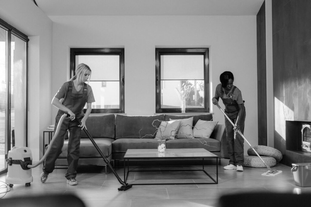 Crafting Captivating Headlines: Your awesome post title goes here Two women in work clothes cleaning a modern living room using a vacuum and mop.