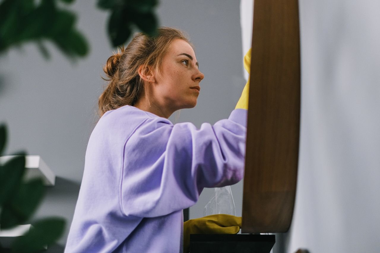 Side view of young focused female in latex gloves standing and cleaning mirror in light room in daytime